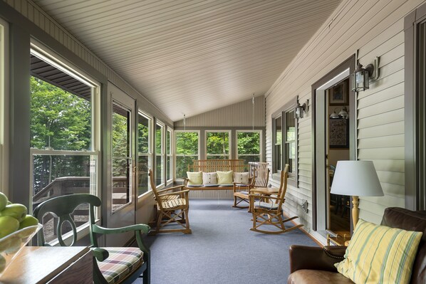 A covered porch with a seating area featuring rocking chairs and a bench, large windows, and a lamp.