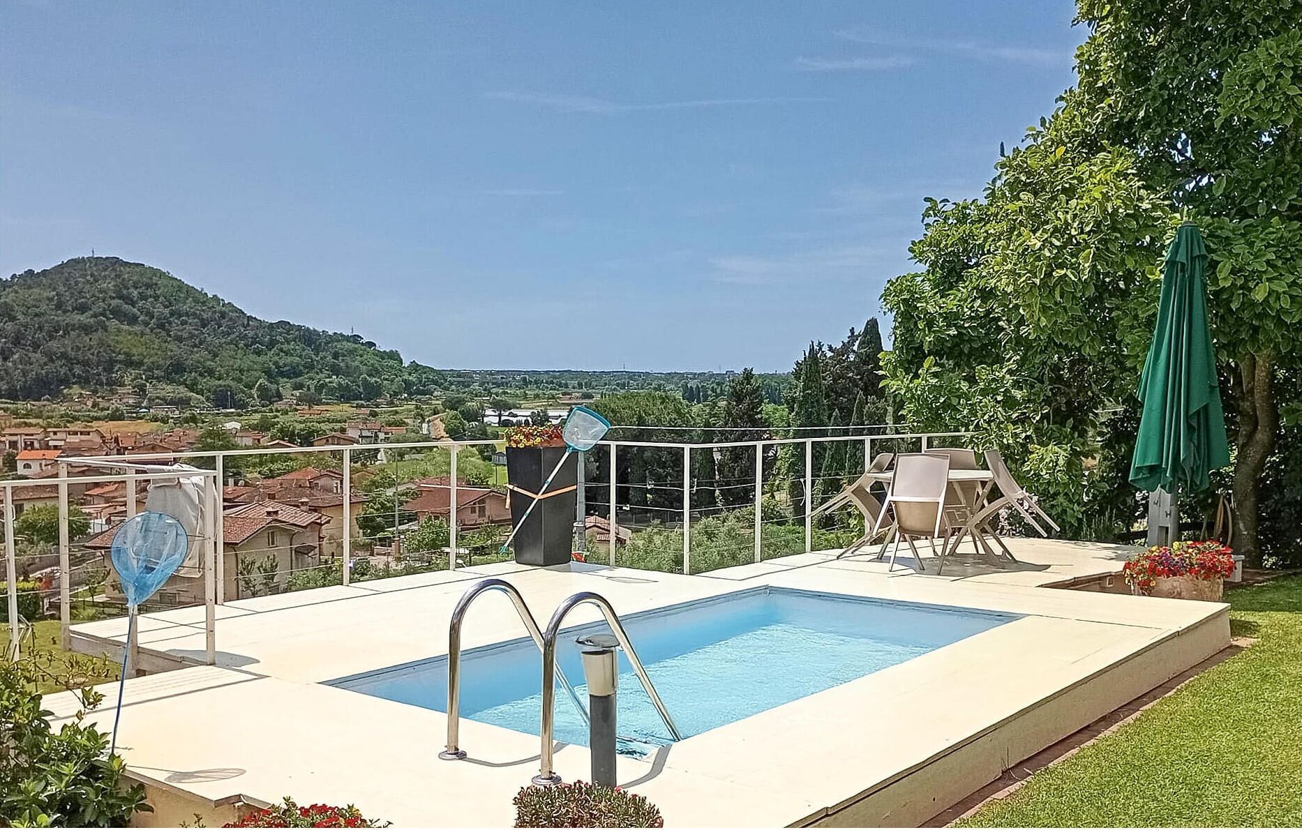 A rectangular pool with a metal ladder, surrounded by a white fence, with a view of a hillside and houses in the background.