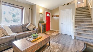 Een woonkamer met een bruine leren bank, een houten salontafel, een rode deur en een trap.