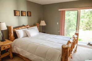 A bedroom with a large bed, wooden headboard, bedside tables, and a view of greenery through a sliding door.
