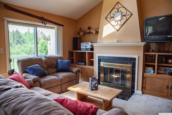 A cozy living room with a fireplace, a television, and a large window offering a view of the outdoors.