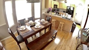 A dining area with a wooden table set for six, a kitchen in the background, and a trash bin to the right.