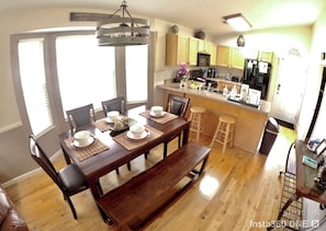 A dining area with a wooden table set for six, a kitchen in the background, and a trash bin to the right.