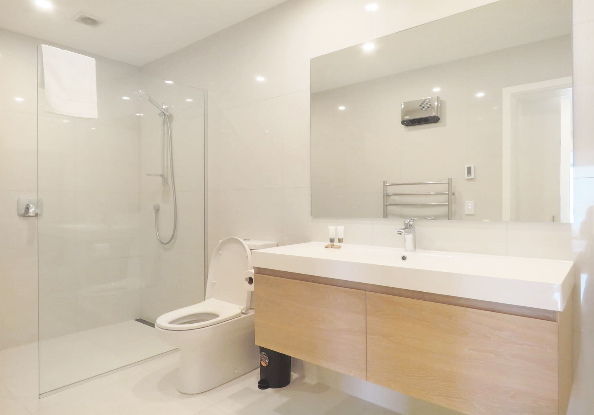 Une salle de bain moderne avec une douche à parois de verre, des toilettes blanches et un meuble-lavabo en bois surmonté d’un miroir.