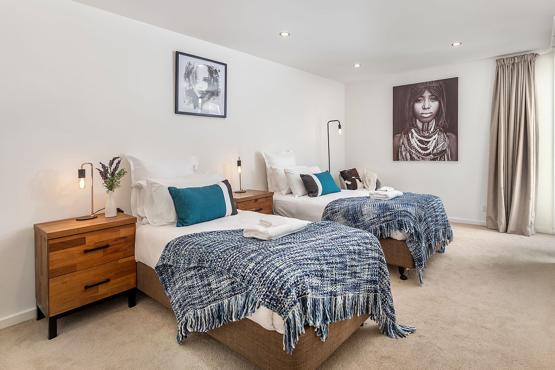 A modern bedroom with two beds, each with a blue and white blanket, white bedding, and a nightstand with a lamp.