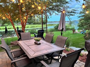 An outdoor patio with a wooden table and chairs, surrounded by greenery and a view of a lake.