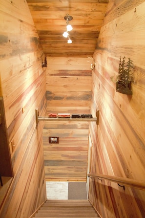 Una escalera de madera con barandilla de metal, una lámpara en el techo y un pequeño árbol decorativo en la pared.