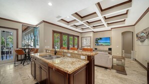A spacious kitchen with granite countertops, a dining area with orange chairs, and a large TV mounted on the wall. - Dog-friendly gated spacious Siesta Key Getaway — Pool, Spa, & Mancave. (Siesta Key)