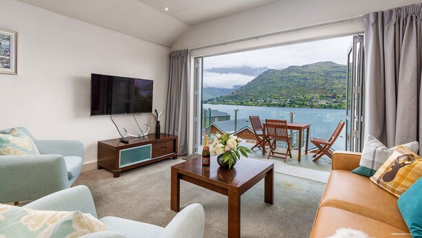 A modern living room with a flat-screen TV, a wooden coffee table with flowers, a sofa, and a dining area with chairs. - Alpine Village - 2 Bedroom Executive Apartment (Queenstown)
