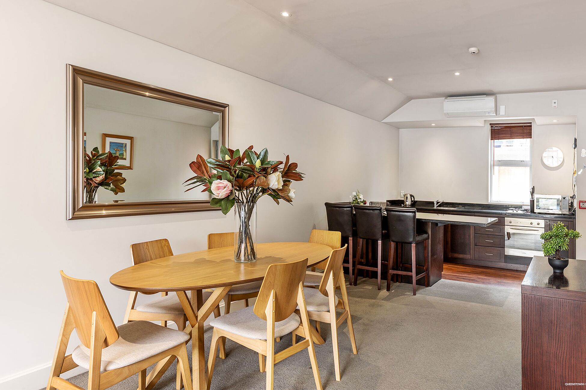 Una cocina moderna con una isla central, un comedor con mesa y sillas de madera, y un espejo grande en la pared.