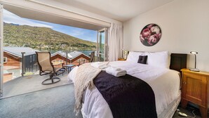 A hotel room with a bed, a nightstand, a lamp, and a view of a lake and mountains. - Alpine Village - 2 Bedroom Executive Apartment (Queenstown)