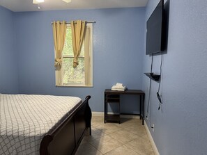 A bedroom with a bed, a window with curtains, a TV mounted on the wall, and a small table with a towel.
