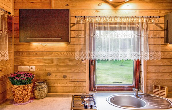 A wooden cabin interior with a gas stove, sink, lace window curtain, and a view of the outdoors.