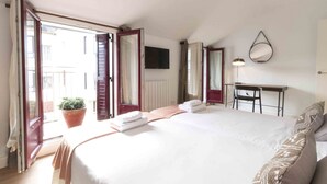 A hotel room with a large bed, a desk with a chair, a potted plant, and a view of the outside. - Atlantic | Rentals Collection (San Sebastián)
