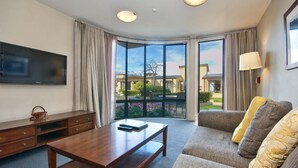 A hotel room with a large TV, a wooden coffee table, a sofa with cushions, and a balcony with a view of other buildings and greenery. - The Beacon - 2 Bedroom Town Center Apartment (Queenstown)
