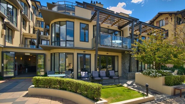 A modern multi-story building with a glass facade, outdoor seating area, and a landscaped garden. - The Beacon - 2 Bedroom Town Center Apartment (Queenstown)