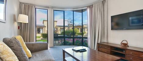 A hotel room with a sofa, a wooden coffee table, a TV, and a large window with curtains.