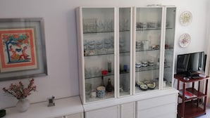 A room with a white cabinet containing glassware and dishes, a framed picture on the wall, and a small table with a vase of flowers. - Les Dunes 2º Solo Familias! (Xeraco Playa)