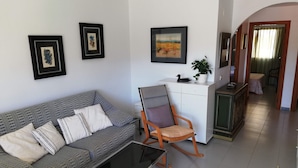 A living room with a grey patterned sofa, a rocking chair, a small table, and a white cabinet. There are framed pictures on the walls and a plant on the cabinet. - Les Dunes 2º Solo Familias! (Xeraco Playa)