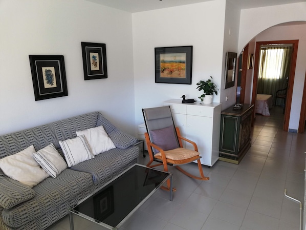 A living room with a grey patterned sofa, a rocking chair, a small table, and a white cabinet. There are framed pictures on the walls and a plant on the cabinet. - Les Dunes 2º Solo Familias! (Xeraco Playa)
