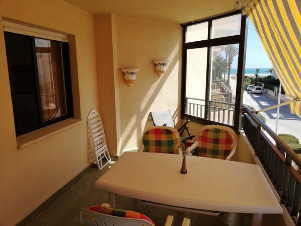A balcony with a table, chairs, a hanging light fixture, and a view of the street and beach.