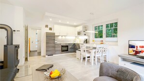 A modern kitchen with white cabinets, a dining area with white chairs, and a large window with a view of trees. - Reetdachhaus Waldidylle (Zirchow/Usedom)