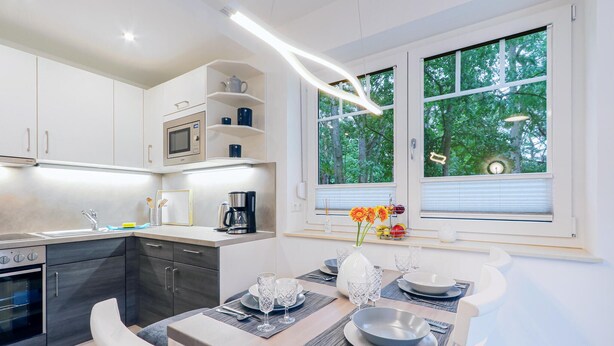 A modern kitchen with white cabinetry, a built-in microwave, and a coffee maker. There is a dining area with a table set for two, featuring clear glassware and a vase with flowers. - Reetdachhaus Waldidylle (Zirchow/Usedom)