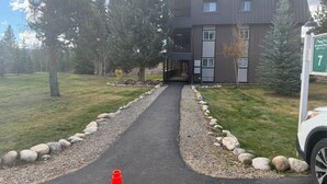 A modern building with a paved driveway leading to a covered entrance, surrounded by trees and a gravel path. - Peaceful cozy place on the river with great views (Winter Park)