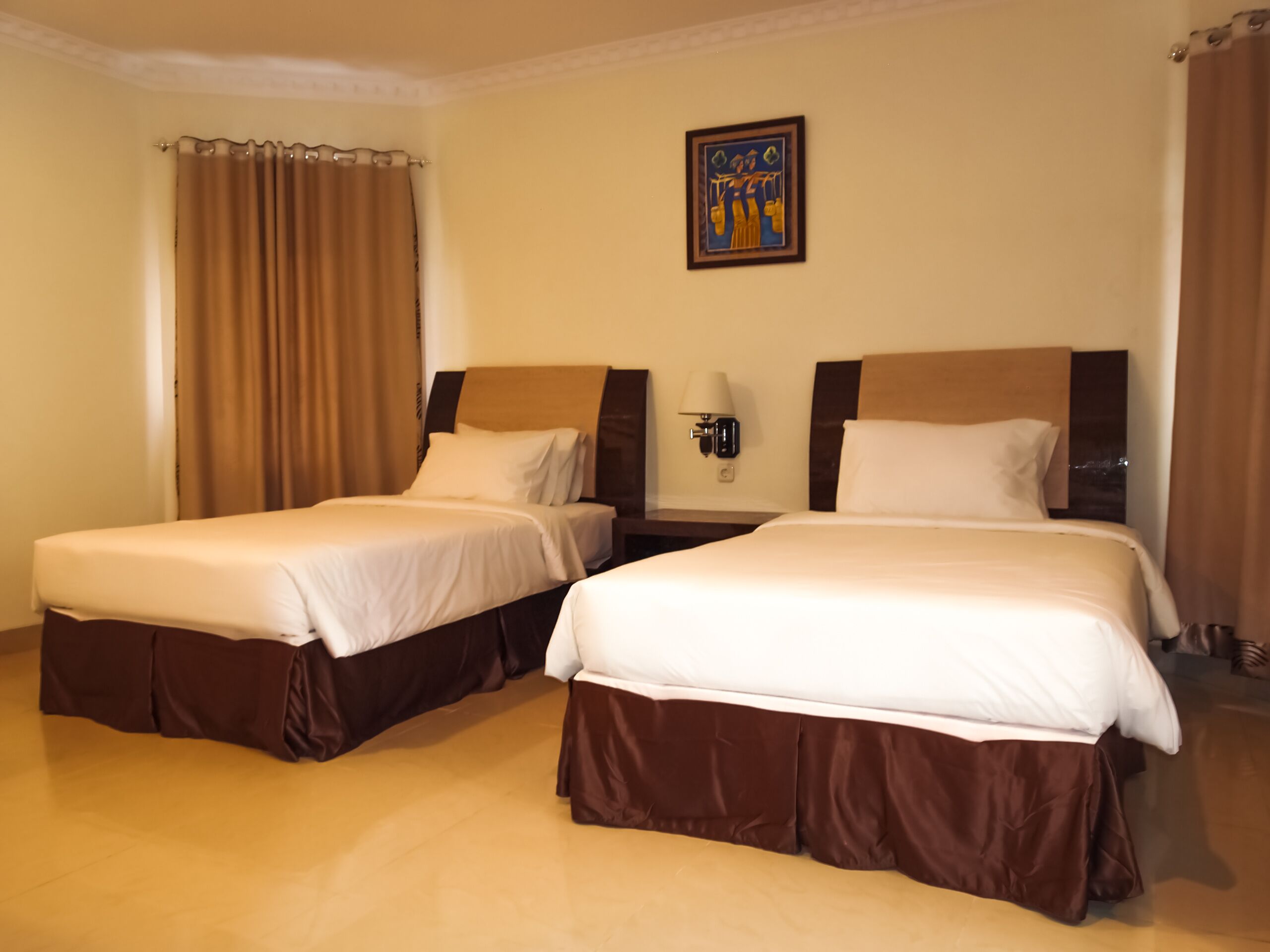 A hotel room with two beds, beige walls, a framed picture, and a nightstand with a lamp.