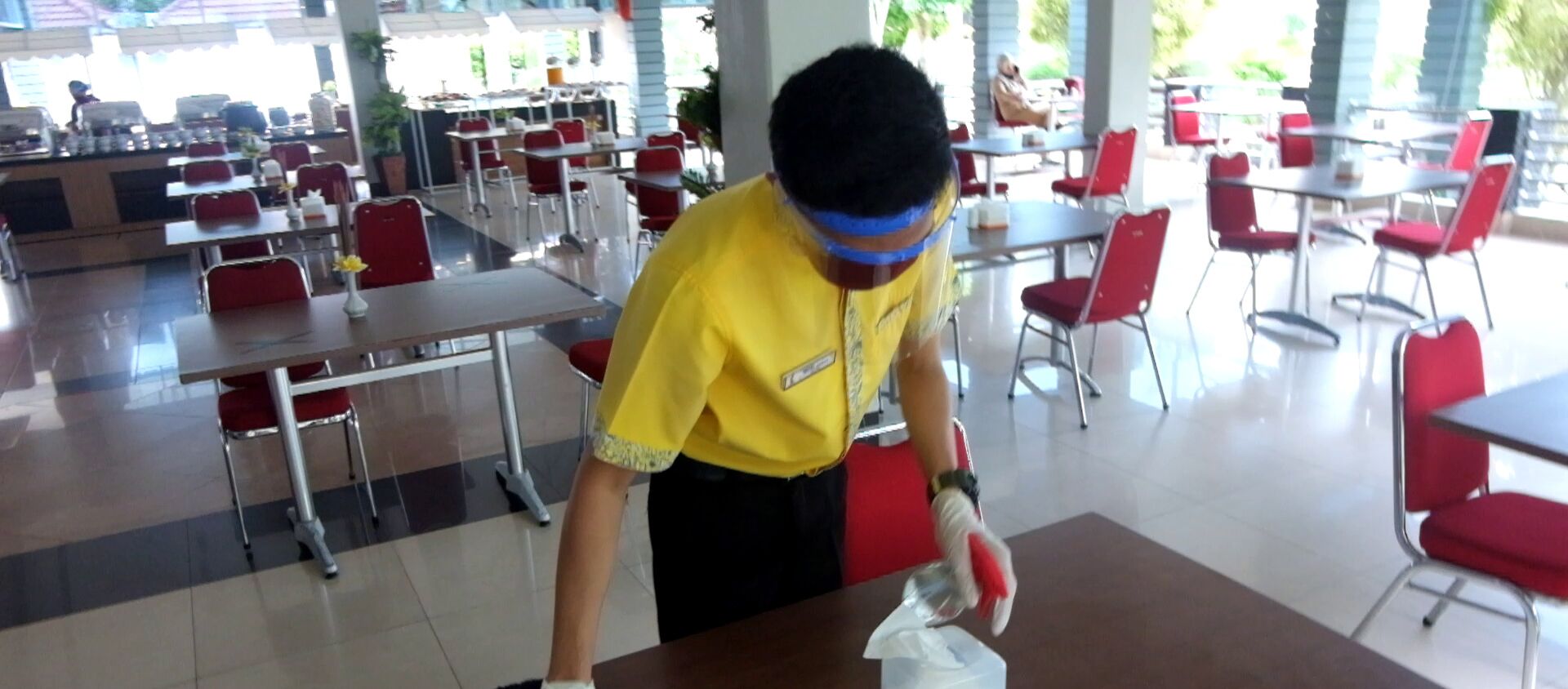 Una cafetería con sillas y mesas rojas, una persona con camisa amarilla limpiando una mesa, y un mostrador con alimentos al fondo.