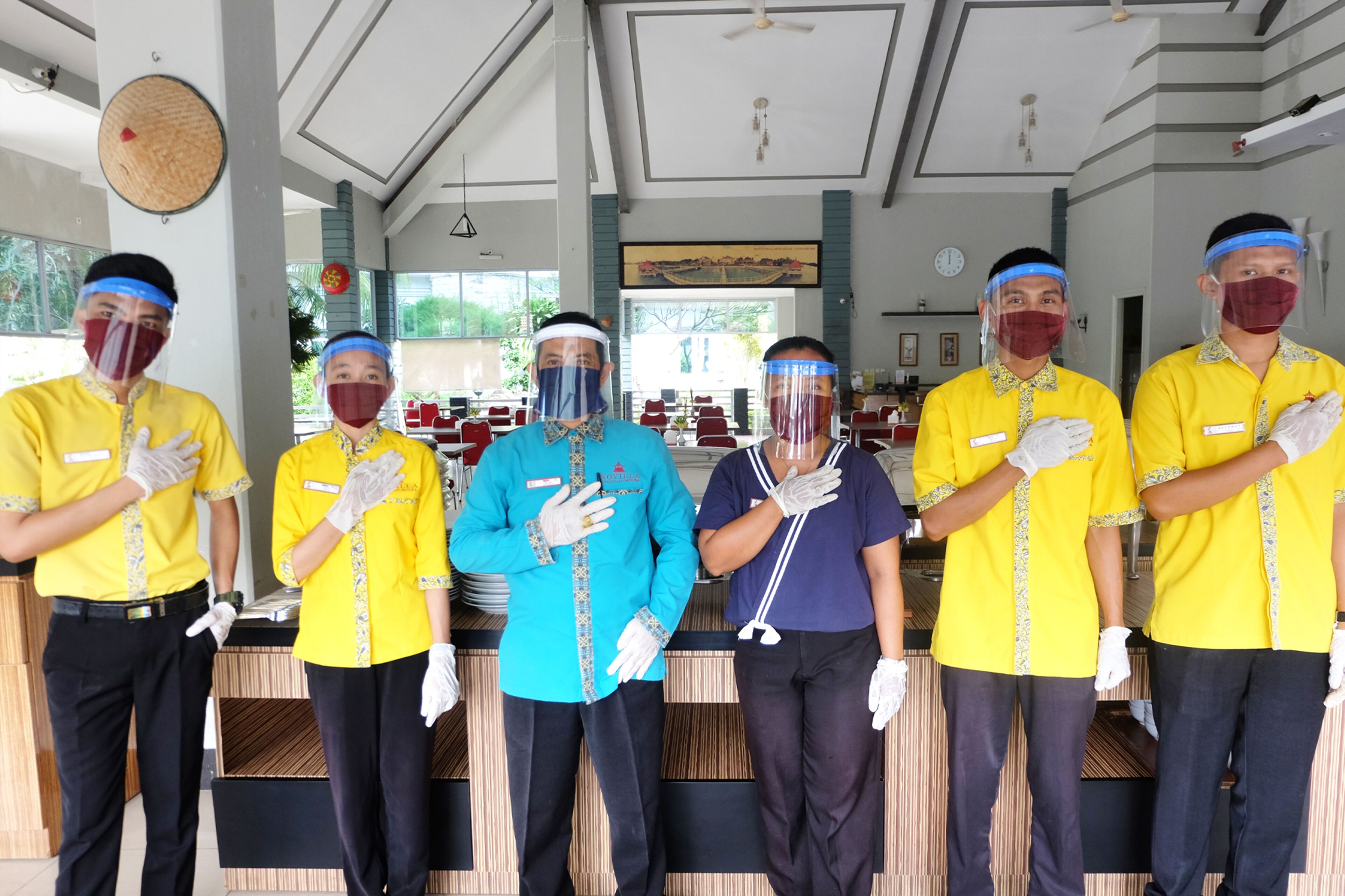 Een groep hotelmedewerkers in gele uniformen staat in een lobby met een receptiebalie, een klok en een groot raam met uitzicht op groen.