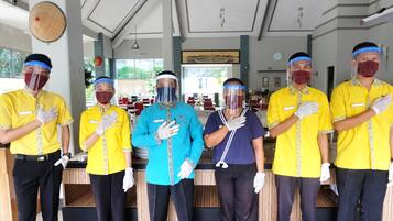 Een groep hotelmedewerkers in gele uniformen staat in een lobby met een receptiebalie, een klok en een groot raam met uitzicht op groen.