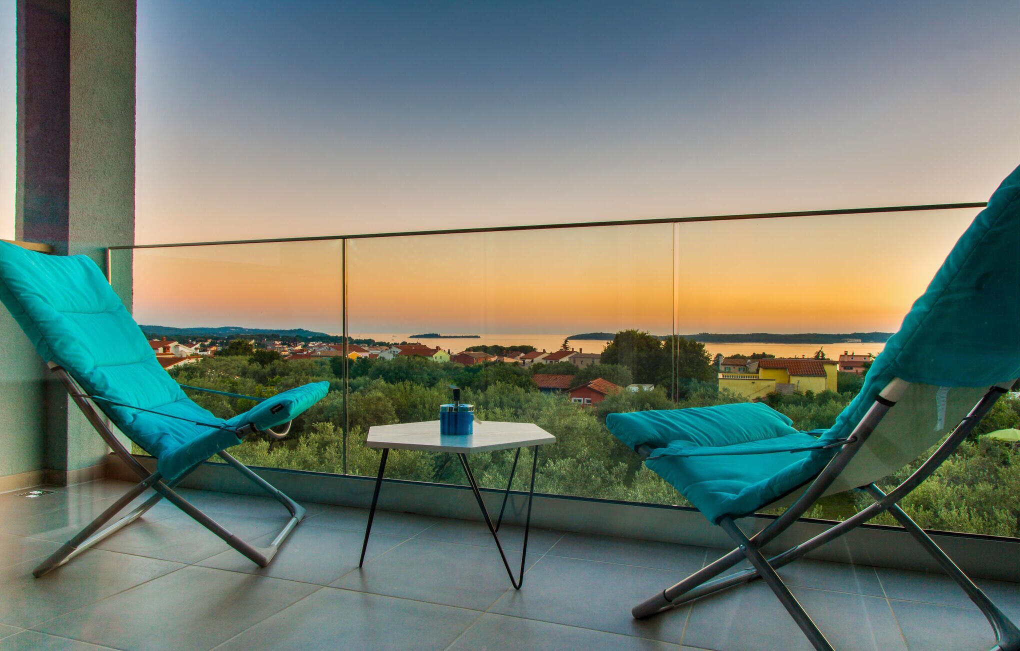 Un balcon avec deux chaises turquoise et une petite table, offrant une vue panoramique sur une ville et l’eau au coucher du soleil.