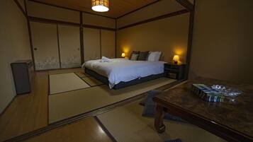 Une chambre de style japonais traditionnel, avec un sol recouvert de tatamis, un lit avec une literie blanche, deux lampes de chevet et une table en bois sur laquelle se trouvent une boîte et un objet en plastique.