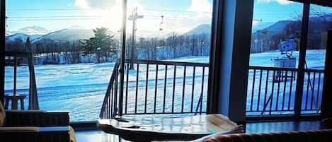 A room with a large window overlooking a snowy landscape, a balcony with a railing, and a seating area with a table and chairs.