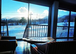 A room with a large window overlooking a snowy landscape, a balcony with a railing, and a seating area with a table and chairs.