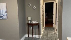 Un pasillo con paredes grises, una mesa de madera oscura con una lámpara y un cartel que dice 'RELAX', y un piso de baldosas.