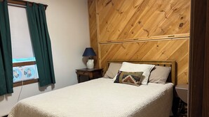 A bed with a quilted cover, a bedside table with a lamp, a window with curtains, and a wooden wall panel. - Woody Creek On-Mountain Ski House - 6 Bedroom, Sleeps 10, Pool Table (Carrabassett Valley)