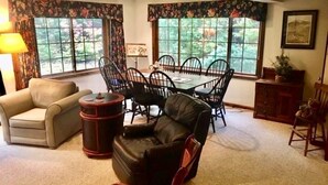 A living room with a sofa, a chair, a dining table with chairs, and a window with floral curtains. - Woody Creek On-Mountain Ski House - 6 Bedroom, Sleeps 10, Pool Table (Carrabassett Valley)