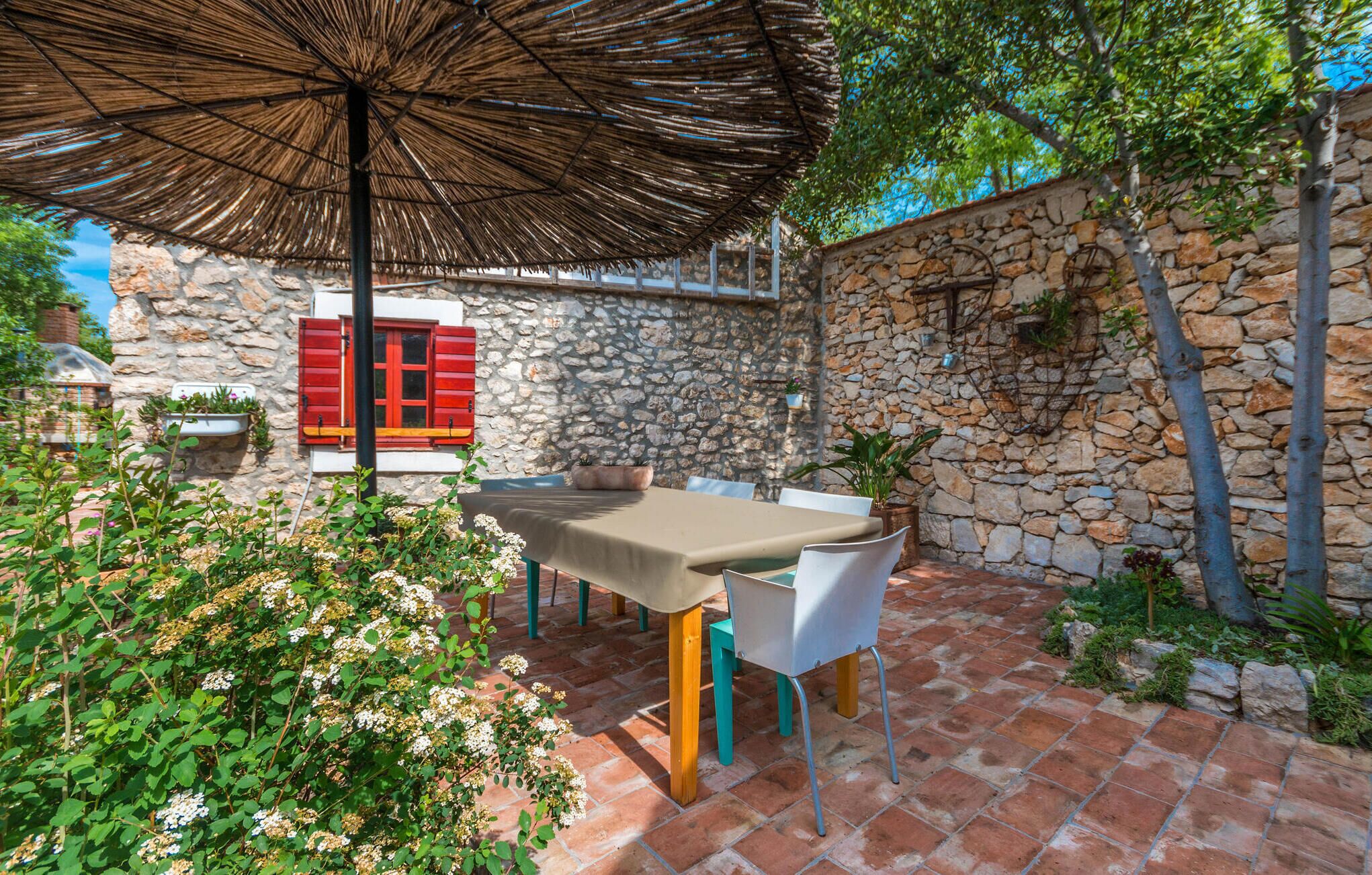 An outdoor patio with a table and chairs, a thatched umbrella, stone walls, and potted plants.