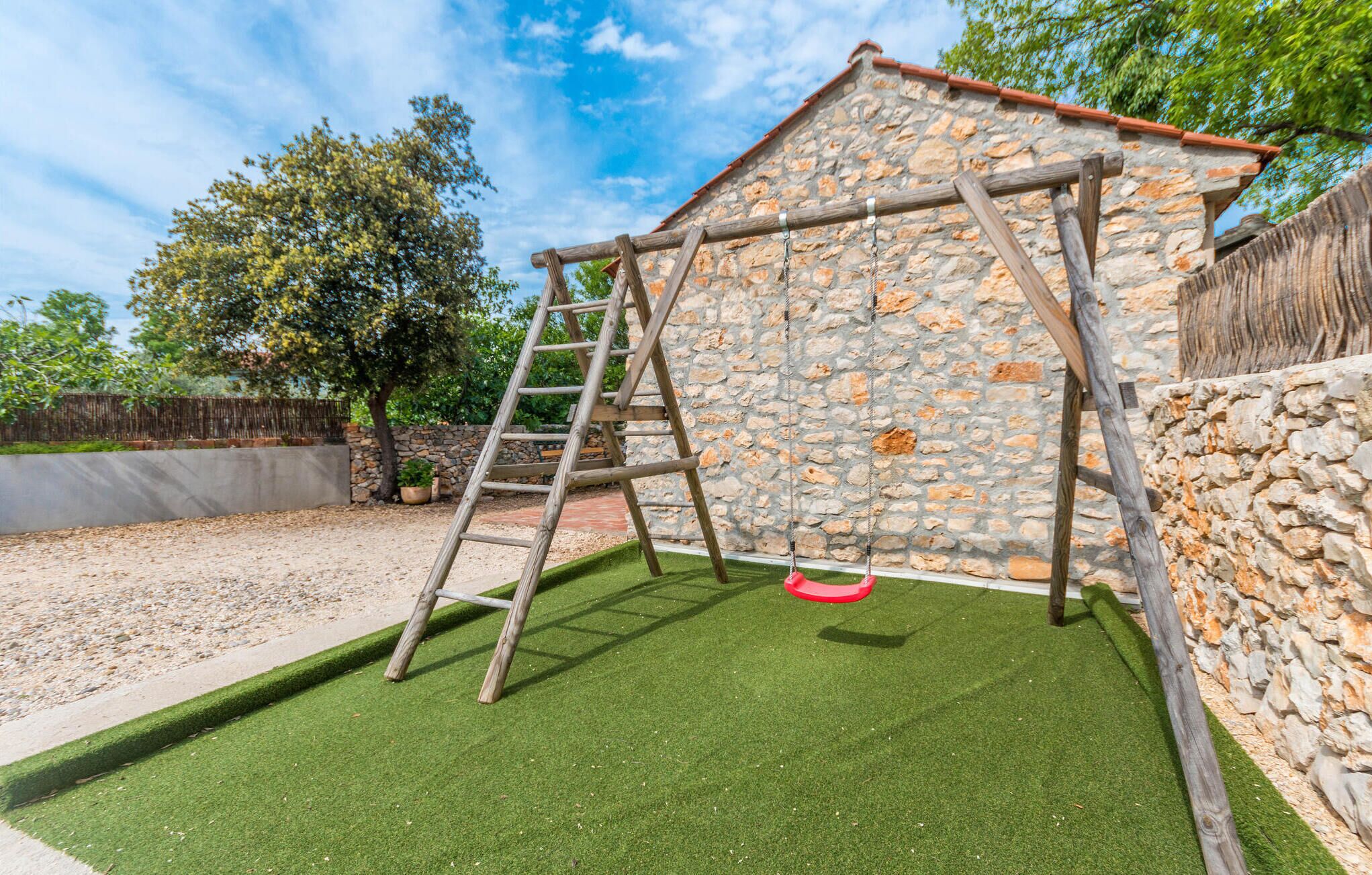Un conjunto de columpios de madera con un muro de piedra de fondo, una escalera y césped artificial verde.