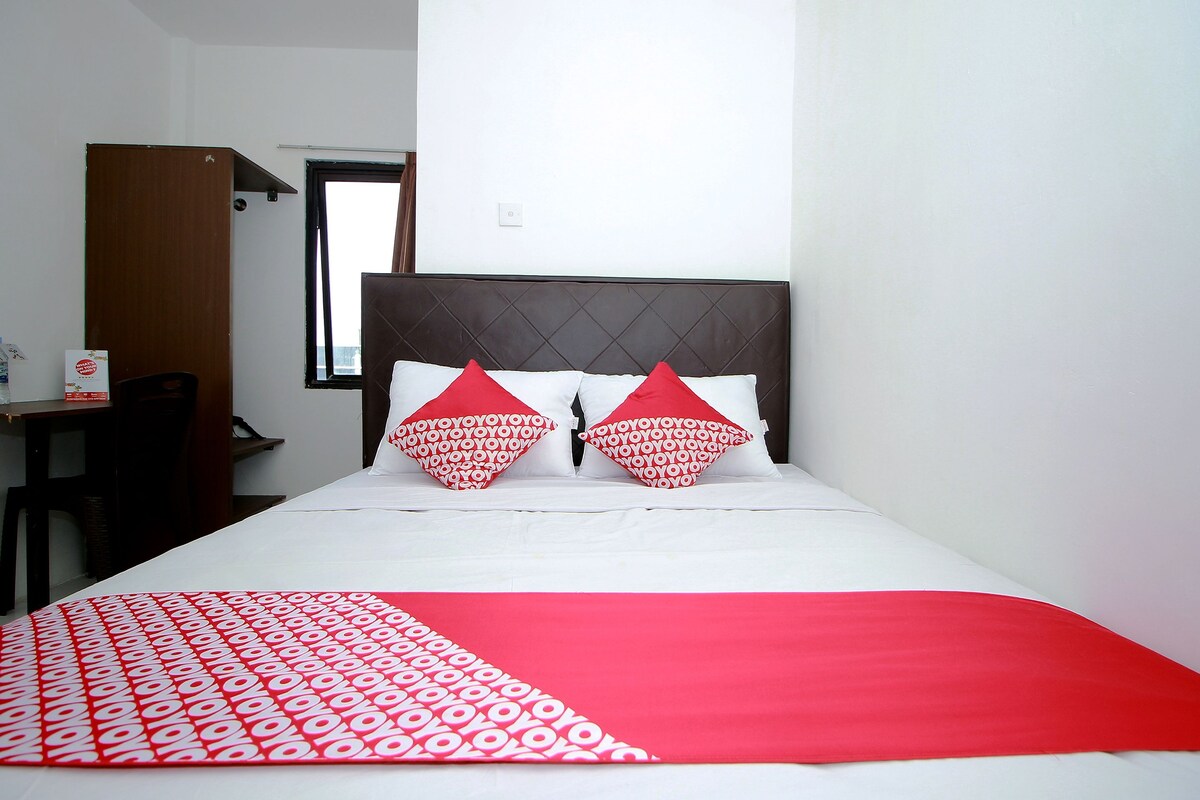 A hotel room with a bed, two pillows, a red and white blanket, a wooden headboard, and a window.