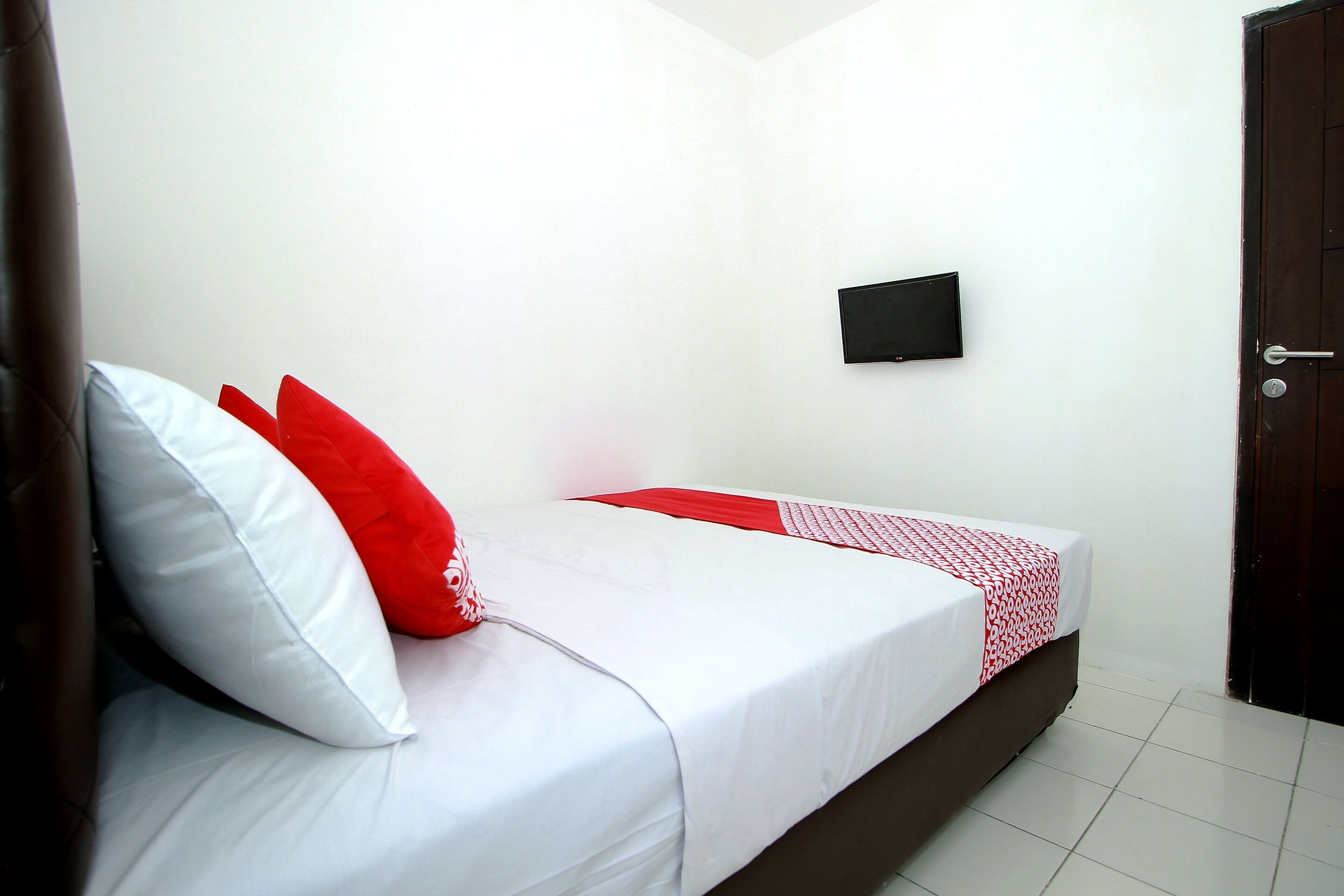 A single bed with white and red bedding, a small black television on the wall, and a wooden door.