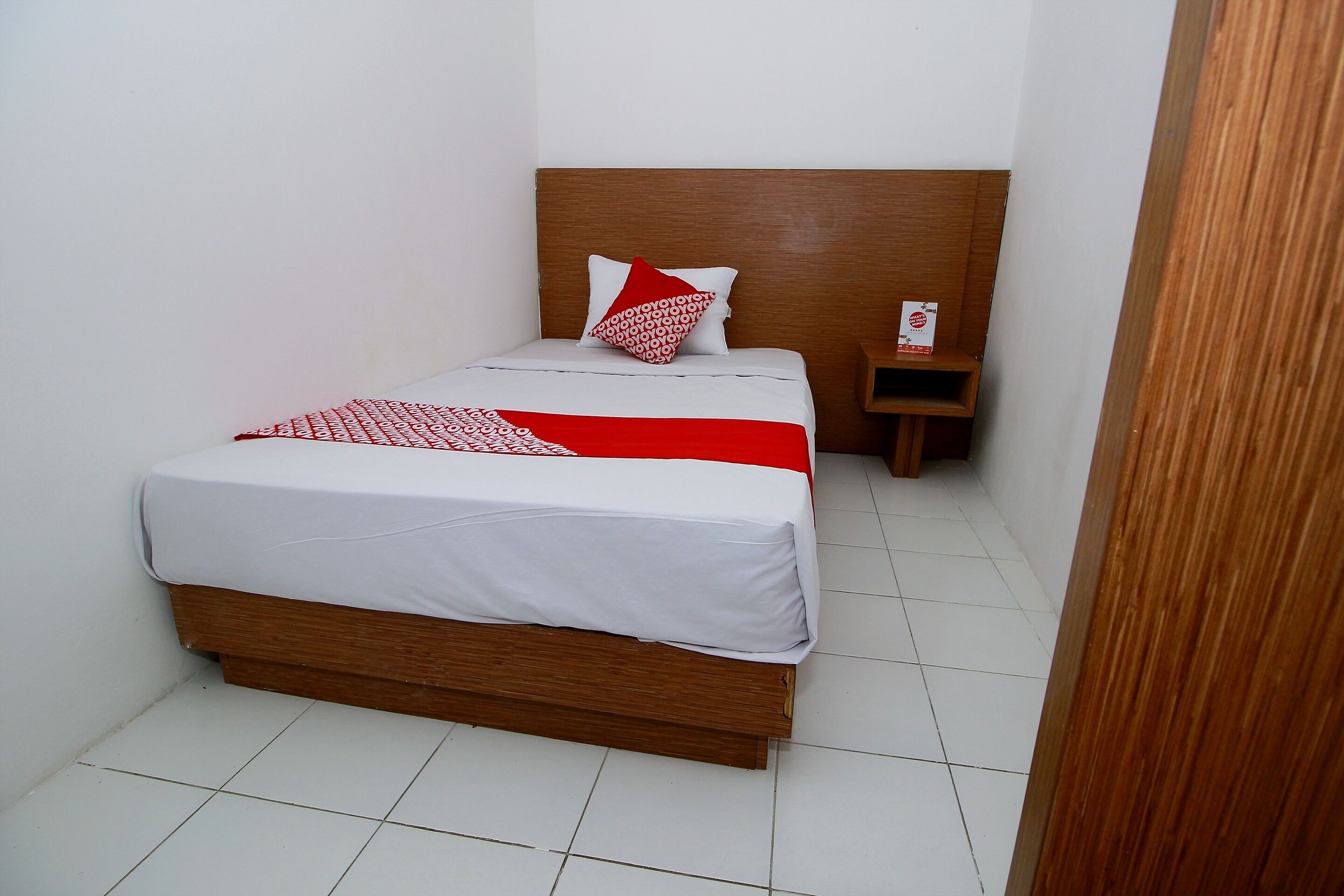 Una cama bien tendida con un cabecero de madera, una mesita de noche pequeña y un cojín estampado en rojo y blanco.