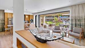 A modern hotel room with a wooden dining table, a sofa, and a view of the outdoor pool and garden.