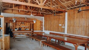 Un interior rústico de madera con varias mesas y bancos, un ventilador de techo y una zona de bar con estantes y botellas.