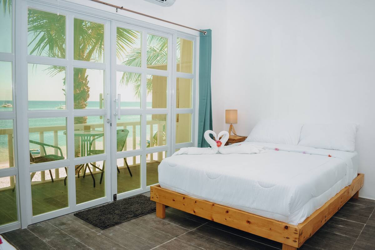 Un dormitorio con una cama de madera, vista a la playa a través de amplios ventanales y una mesita con una lámpara.