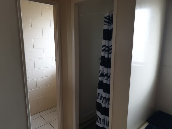 Un pasillo estrecho con una pared de ladrillo blanco, una puerta que da a un baño con cortina de ducha a rayas y un pequeño banco azul.