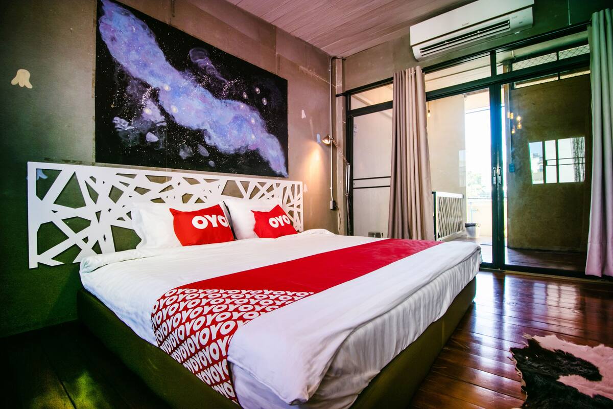 A large bed with a white headboard and red pillows, a red and white patterned blanket, and a galaxy-themed painting on the wall.