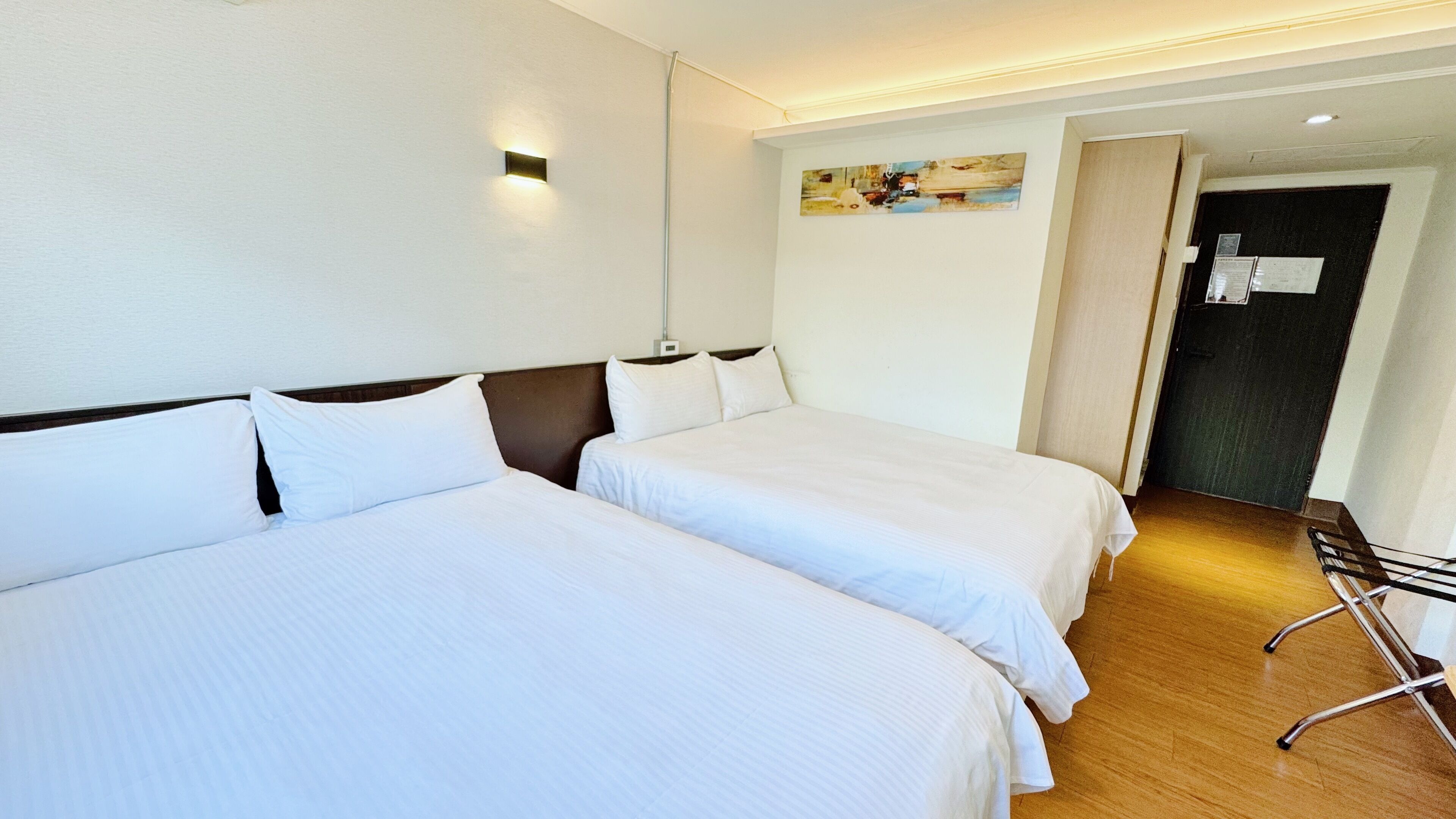 A hotel room with two single beds, a wooden floor, a painting on the wall, and a door with a sign.